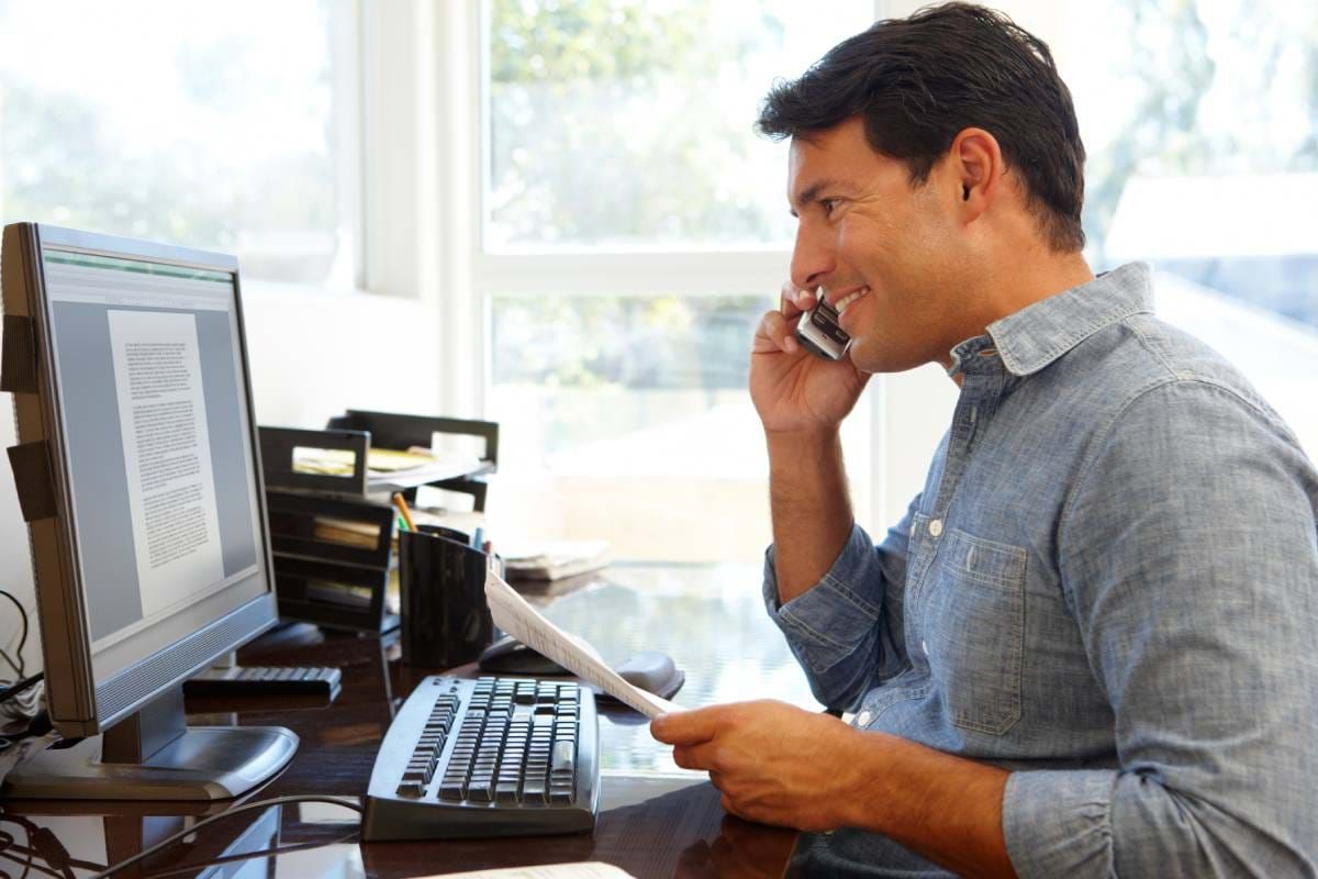 A man working from home in front of his computer while talking on the phone