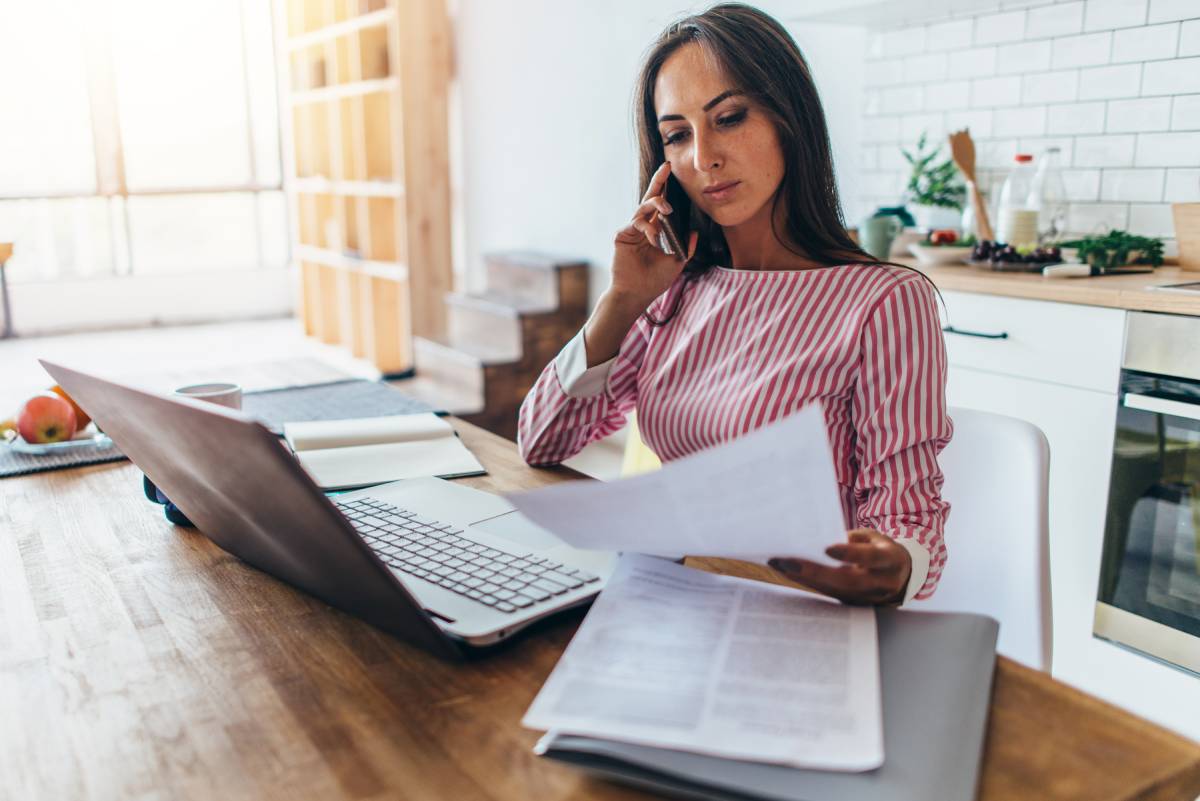 A woman wearing a red and white striped top talking on the phone, and using a laptop while working remotely