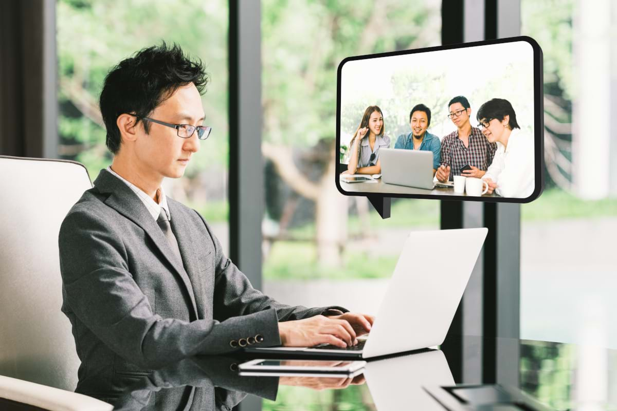 A man in a suit with glasses sitting at a table, video conferencing on his laptop with four other people 